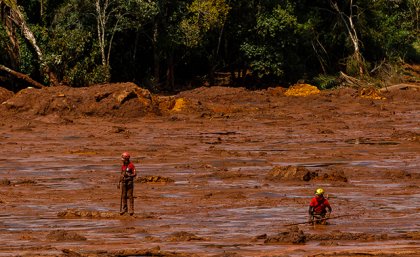 Two people in red shirts and helmets stand in red mud, there are trees in the background